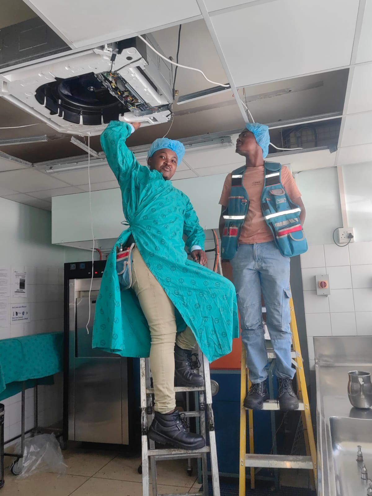 Two technicians in medical PPE servicing a ceiling-mounted air conditioning unit at a hospital